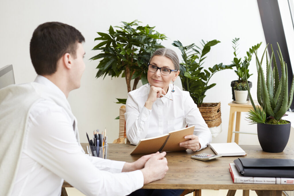 candid shot of attractive positive middle aged female ceo in eyeglasses making notes in copybook, listening to talented prospective young male job candidate during interview in her cozy office